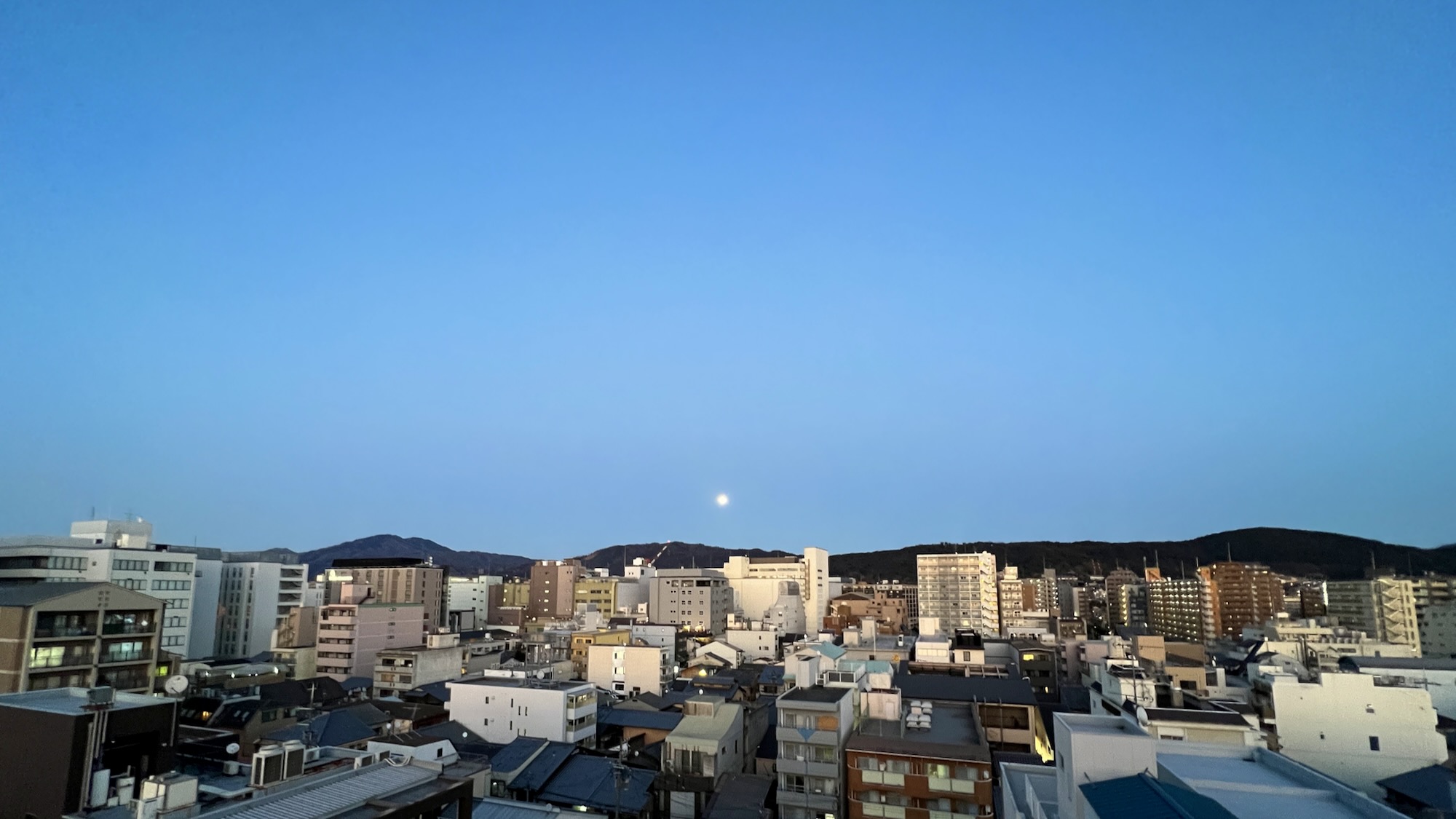 Full moon low over the horizon during a blue twilight sky. Just below are the eastern mountains of Kyoto, and many buildings
